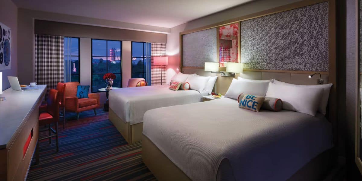 A modern resort hotel room with two queen beds, colorful accent pillows, a desk with a laptop, a sitting area with orange chairs, large windows offering city views at dusk, and stylish striped carpeting.