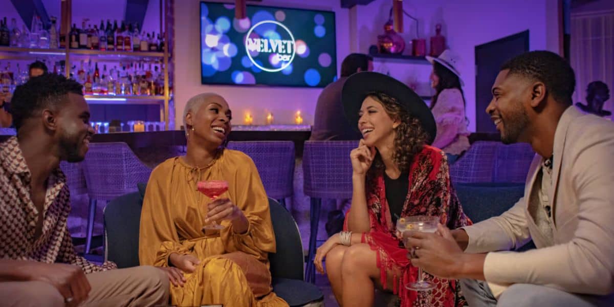 Four friends sit together in a stylish resort lounge, laughing and talking. Two women and two men hold colorful drinks, with a "Velvet" sign glowing on a TV in the background. The room is warmly lit with purple tones.