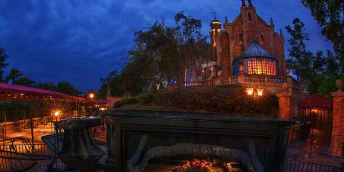 A haunted mansion, part of the Haunted Mansion Update, glows with orange and purple lights behind trees and iron gates at night. A gothic carriage and eerie, dimly lit atmosphere enhance this Save Classic spooky scene.