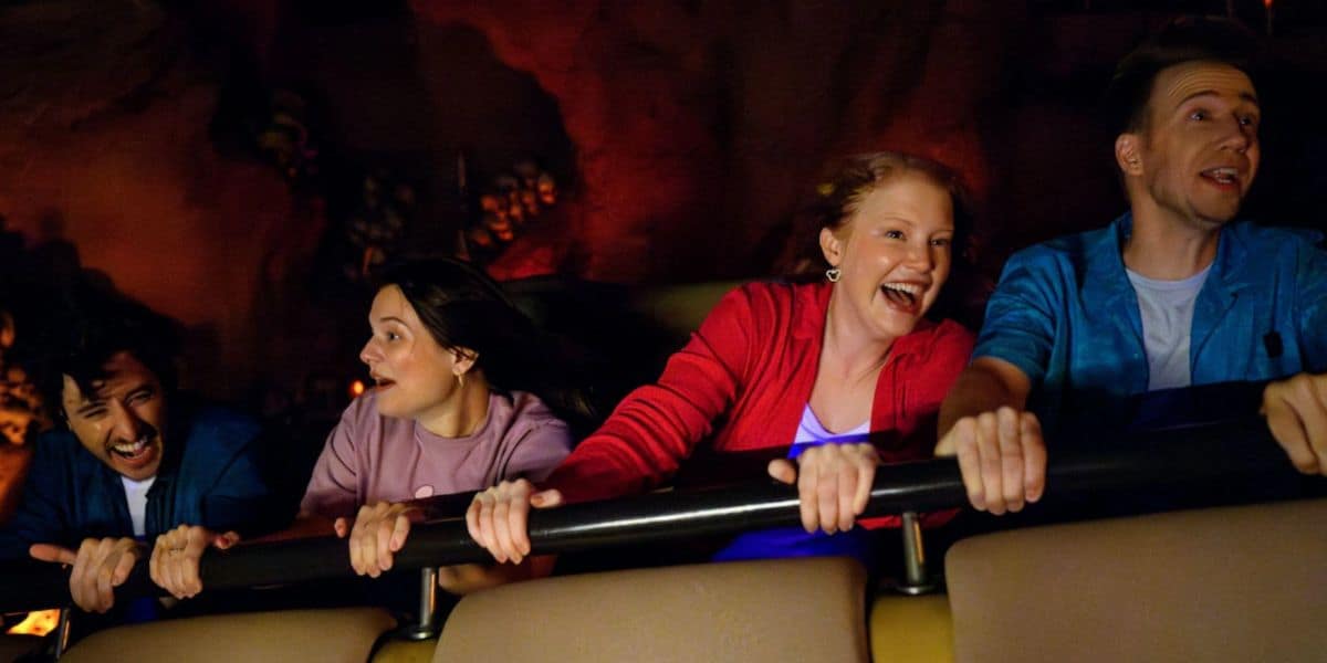 Four people ride a roller coaster, holding onto a safety bar and smiling with excitement as they speed through a dark, Indiana Jones-themed environment.