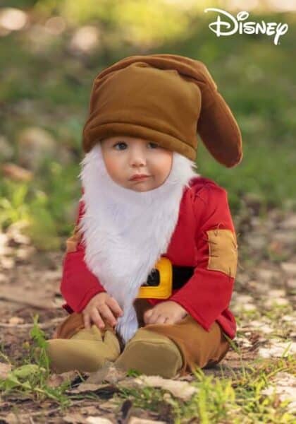 A young child dressed in Disney Costumes as Grumpy Dwarf, wearing a large brown hat, white beard, red shirt, brown pants, and boots, sits outdoors on the ground. The Disney logo appears in the upper right corner.