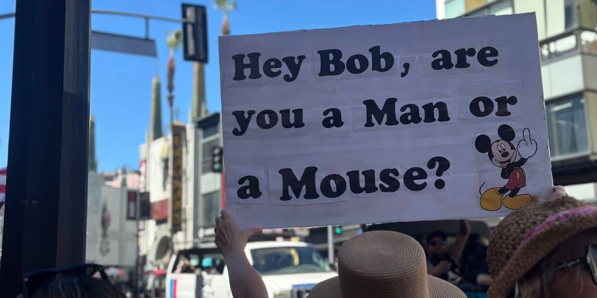 A person holds a sign on a city street that reads, "Hey Bob, are you a Man or a Mouse?" with an image of a cartoon mouse standing and smiling on the right side of the poster.