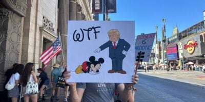 A person holds a sign on a busy street. The sign shows Donald Trump pointing at a sad, kneeling Mickey Mouse with "WTF" written above. An American flag and pedestrians are visible in the background.