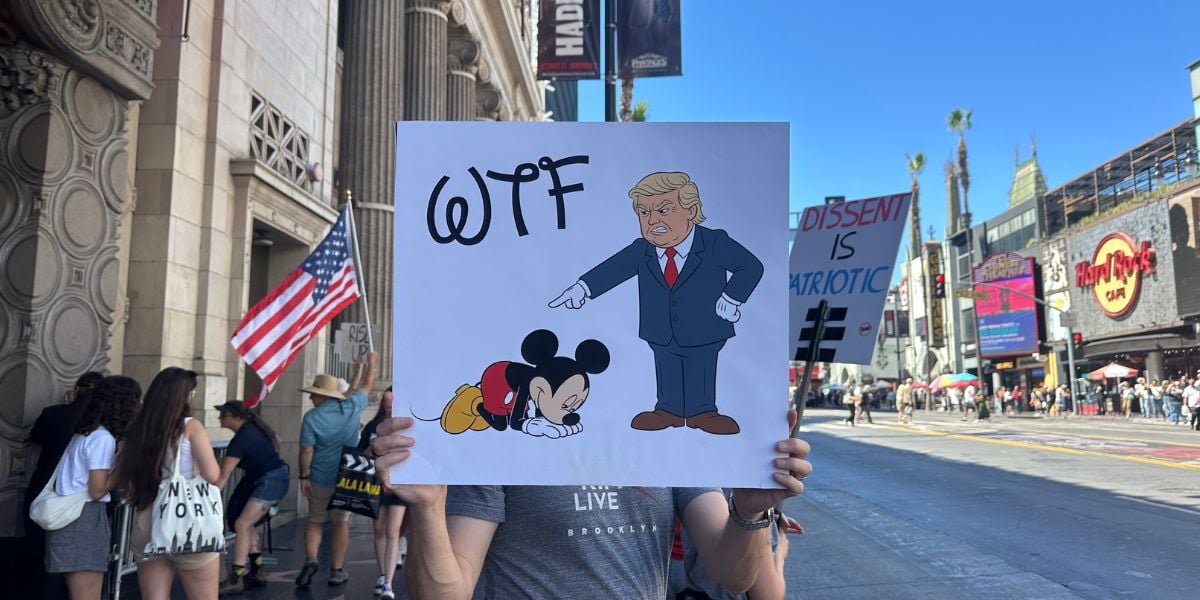 A person holds a sign on a busy street. The sign shows Donald Trump pointing at a sad, kneeling Mickey Mouse with "WTF" written above. An American flag and pedestrians are visible in the background.
