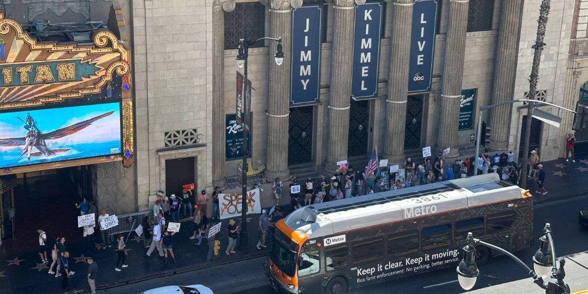A crowd of people holding signs protest outside the Jimmy Kimmel Live studio in Hollywood while a Metro bus passes by on the street. The El Capitan Theatre marquee is visible on the left.