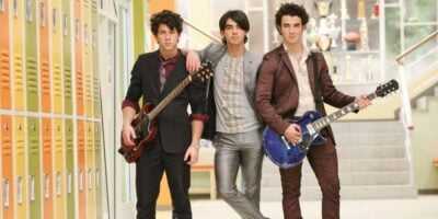Three young men in suits stand in a school hallway with yellow lockers and trophies in the background; two hold electric guitars while the one in the center rests his arm on the other's shoulder, reminiscent of a Disney-era Jonas Brothers reunion.