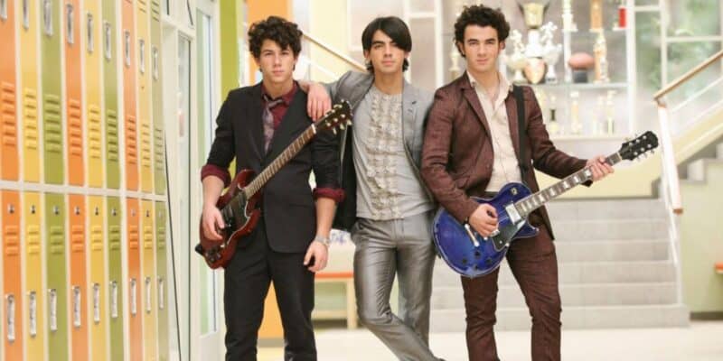 Three young men in suits stand in a school hallway with yellow lockers and trophies in the background; two hold electric guitars while the one in the center rests his arm on the other's shoulder, reminiscent of a Disney-era Jonas Brothers reunion.