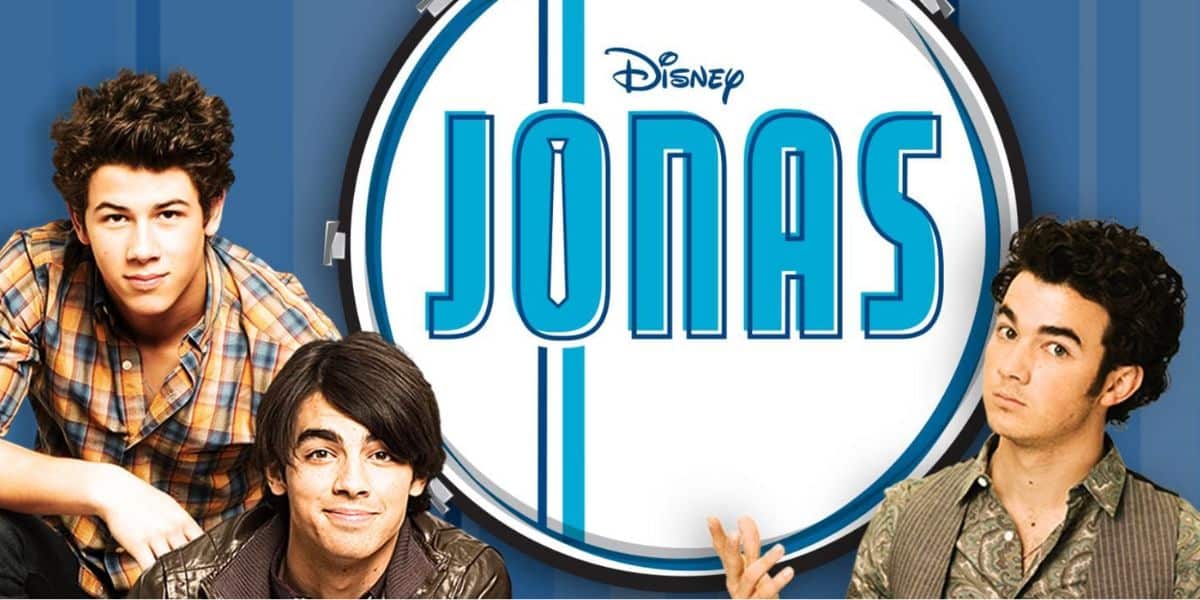 Three young men pose in front of a large drum with the Disney “JONAS” logo, representing the Jonas Brothers. The blue background features vertical stripes, and each is dressed casually with relaxed expressions.
