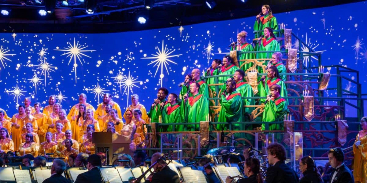 A choir dressed in green and gold robes sings on tiered risers in front of an orchestra, with a blue background featuring large, glowing star designs for a festive Christmas performance.