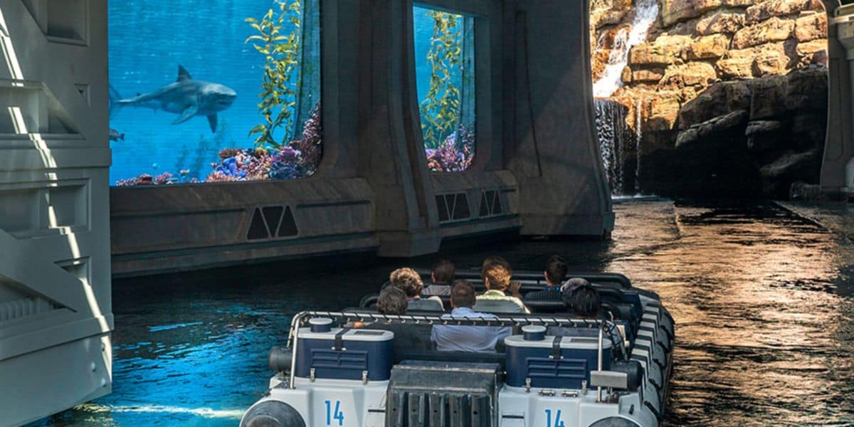 A group of people sit in a jurassic park ride vehicle, passing by large aquarium windows with a shark swimming inside and rocky scenery in the background.