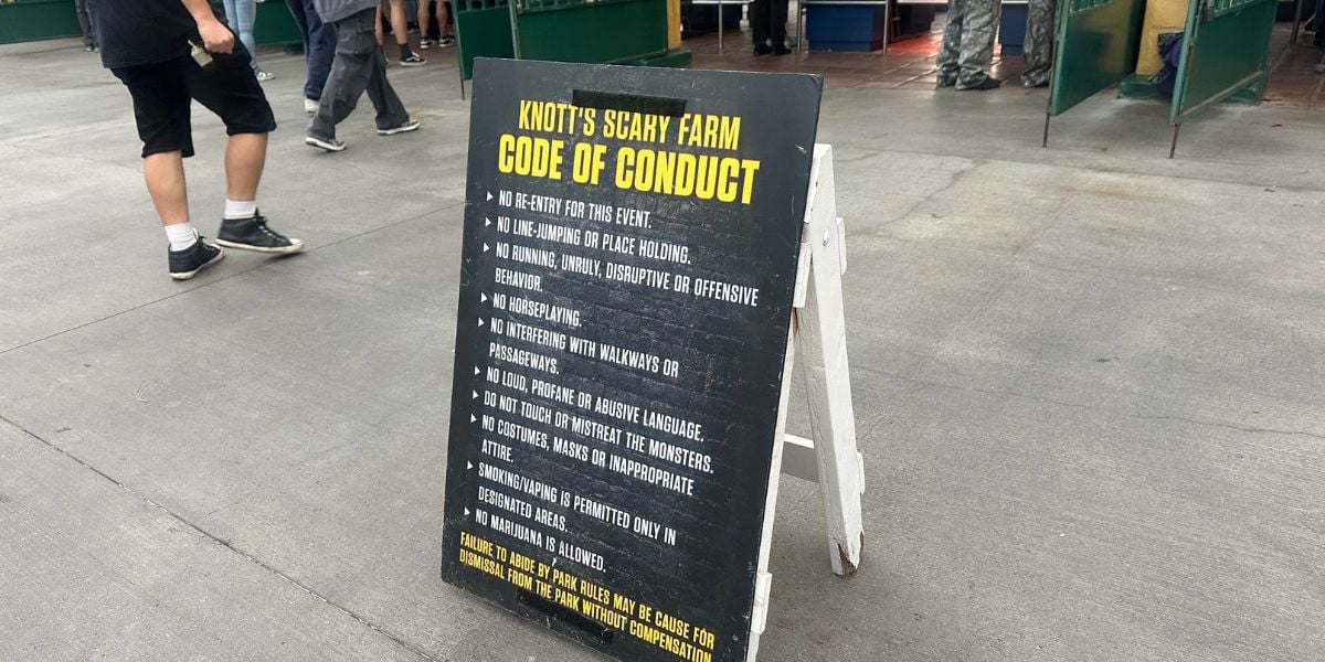 A black sandwich board sign displays the "Knott’s Scary Farm Code of Conduct" with rules in white and yellow text. People walk nearby; the sign is set on a concrete surface near a queue area.