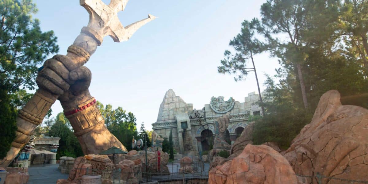 A large stone hand holding a trident rises among rocky structures and trees in Islands of Adventure, with an ancient temple-like building in the background under a clear sky.