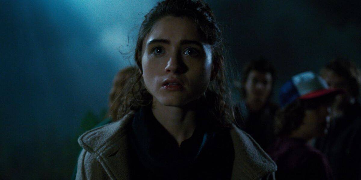 A young woman with wavy brown hair looks ahead with a worried expression at night, surrounded by a group of people in the dim background, illuminated by blue-tinted light, evoking a scene from Stranger Things.