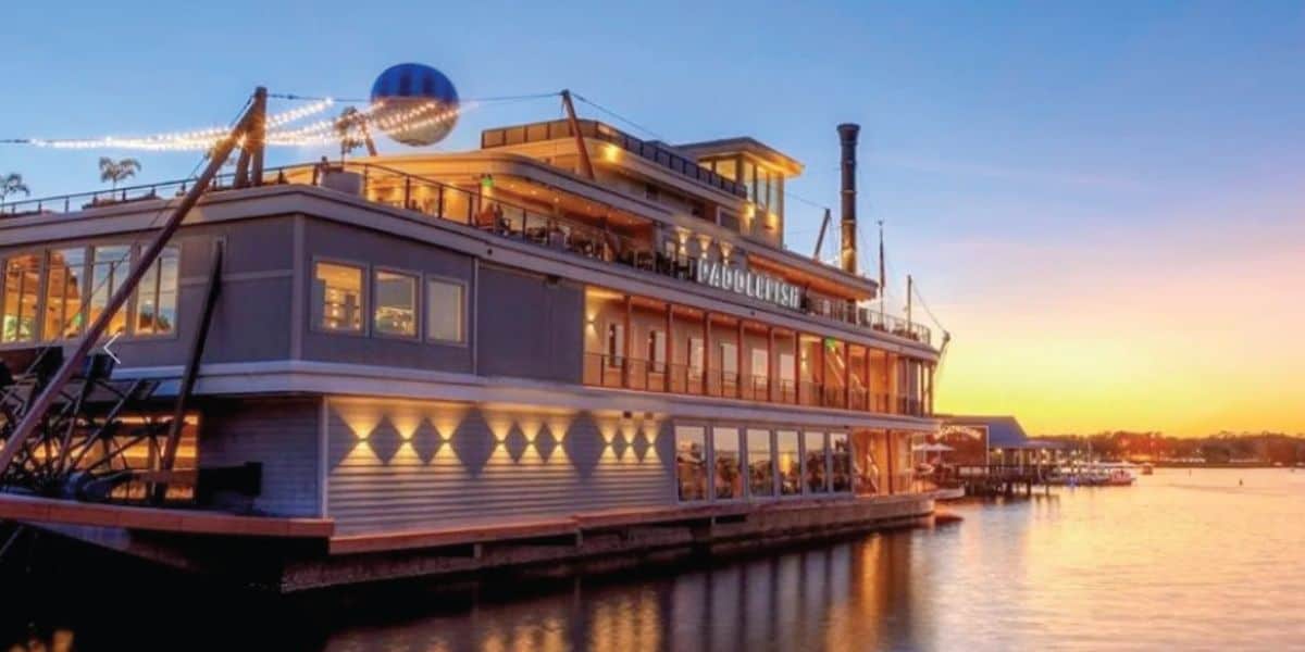 A large, multi-story paddlewheel boat reminiscent of classic Disney riverboats is docked on calm water at sunset, with glowing lights strung above and warm light shining from the windows. The sky is clear, blending from blue to orange near the horizon.