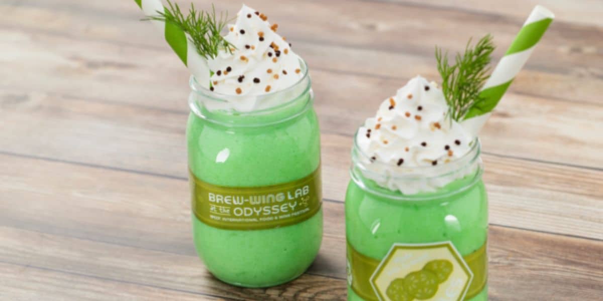 Two green milkshakes in mason jars topped with whipped cream, a sprig of dill, and black seeds evoke a playful Disney vibe. Each jar has a green and white striped straw and a unique label, placed on a wooden surface.
