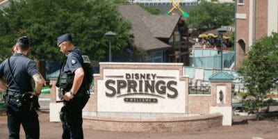 Two police officers stand near the “Disney Springs Welcomes You” entrance sign at Disney World, with buildings, trees, and people visible in the background on a sunny day.