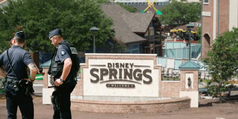 Two police officers stand near the “Disney Springs Welcomes You” entrance sign at Disney World, with buildings, trees, and people visible in the background on a sunny day.