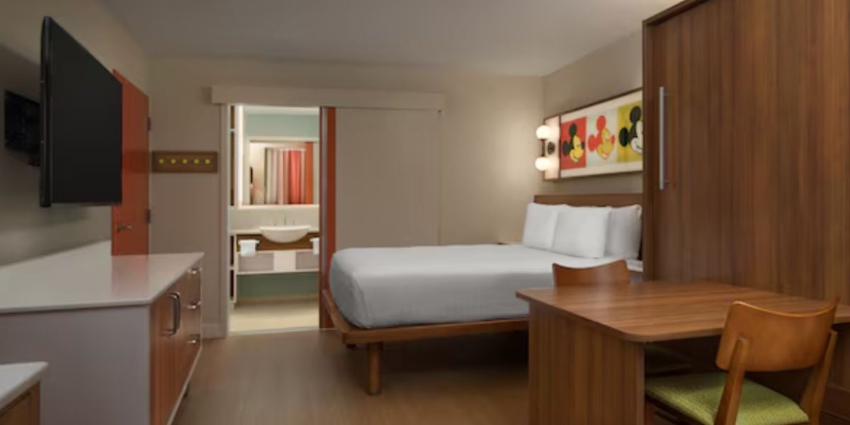 A modern Pop Century hotel room with a bed, wooden desk and chair, wall-mounted TV, and a small kitchenette; a bathroom with sink and mirror is visible through an open sliding door.