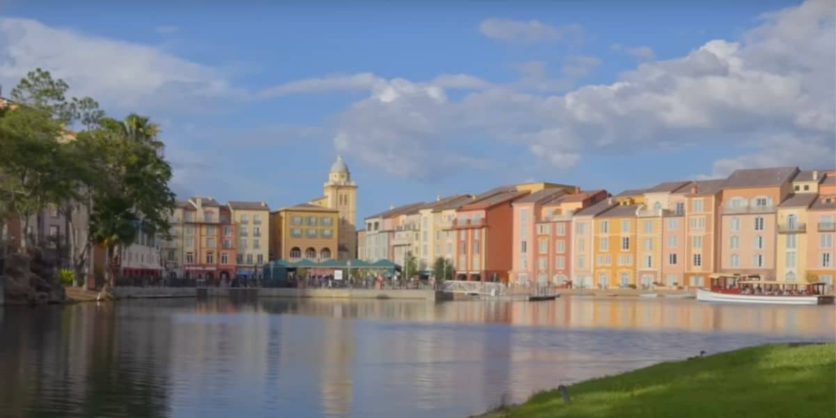 A vibrant waterfront resort scene featuring colorful, Mediterranean-style buildings lining the harbor, a boat docked by the shore, and a clock tower rising in the background under a partly cloudy blue sky.
