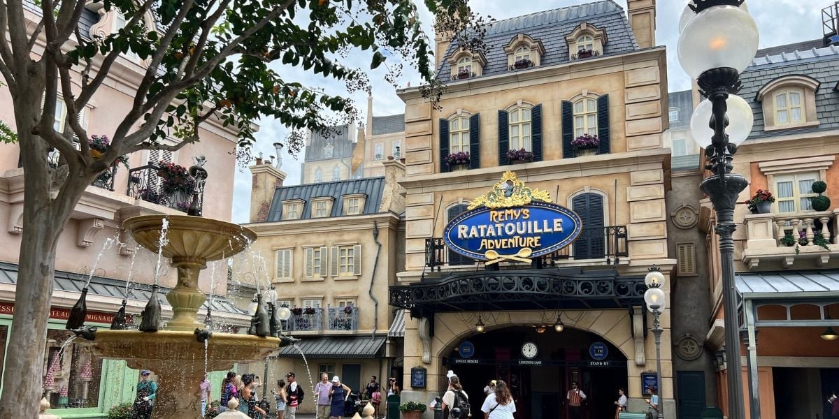 People walk and gather near a fountain in front of the entrance to Remy’s Ratatouille Adventure, a French-themed building with decorative windows and signs, at EPCOT theme park.