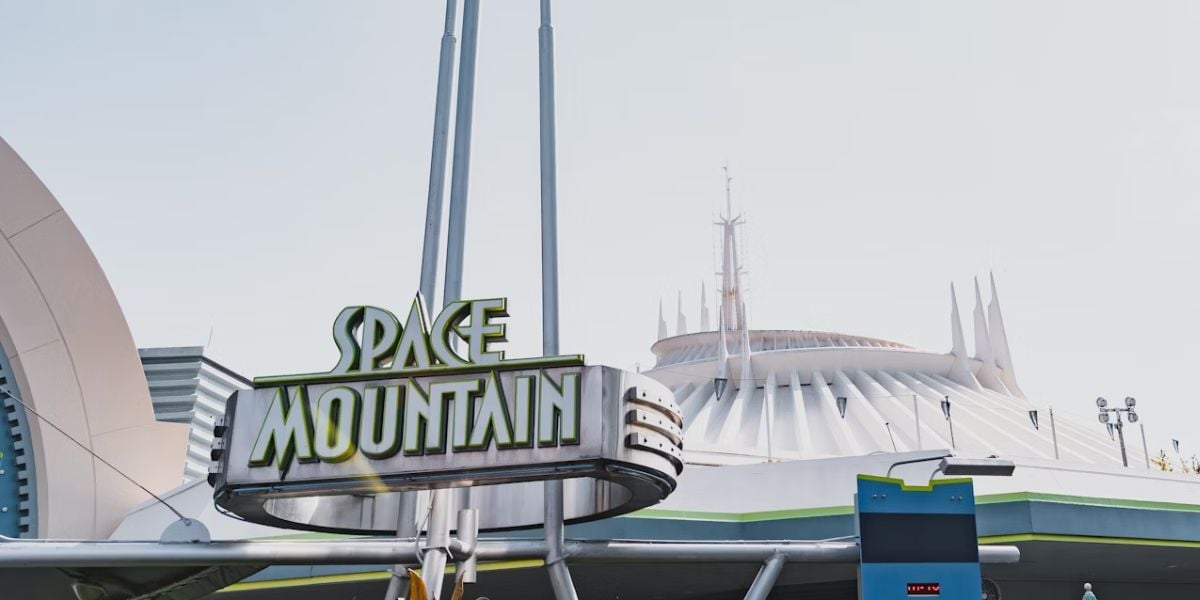 The Space Mountain sign stands in front of the futuristic white dome and spires of the iconic Space Mountain attraction at a Disney theme park under a bright, clear sky.