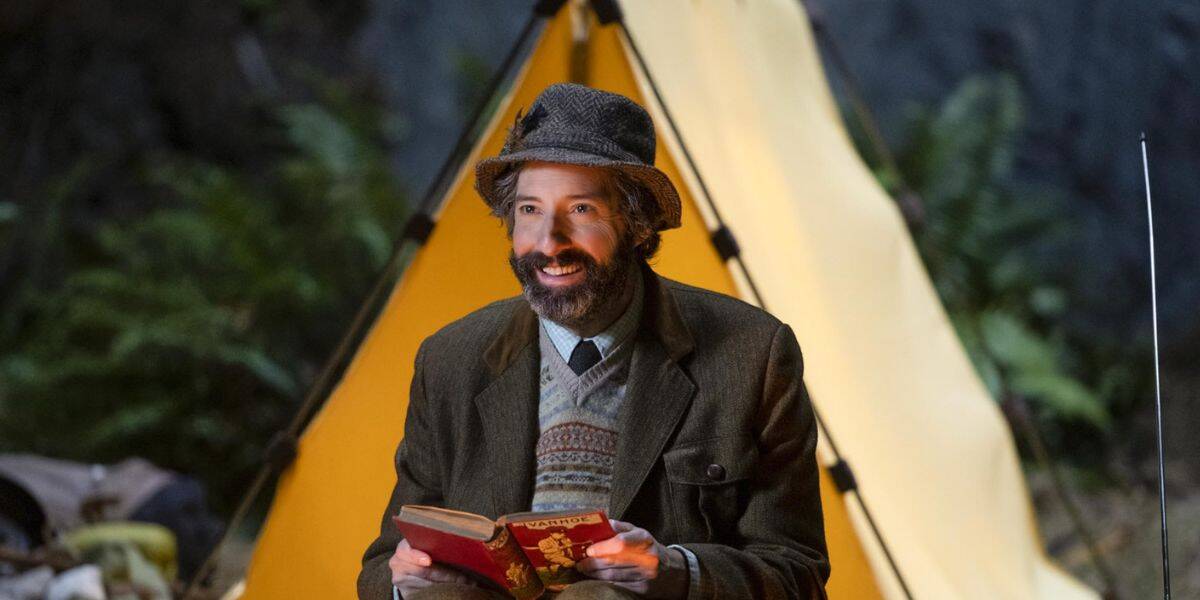 A bearded man in a hat and tweed jacket sits in front of a yellow tent, smiling and holding an open book, surrounded by greenery that feels almost Disney-like in its enchanting outdoor charm.