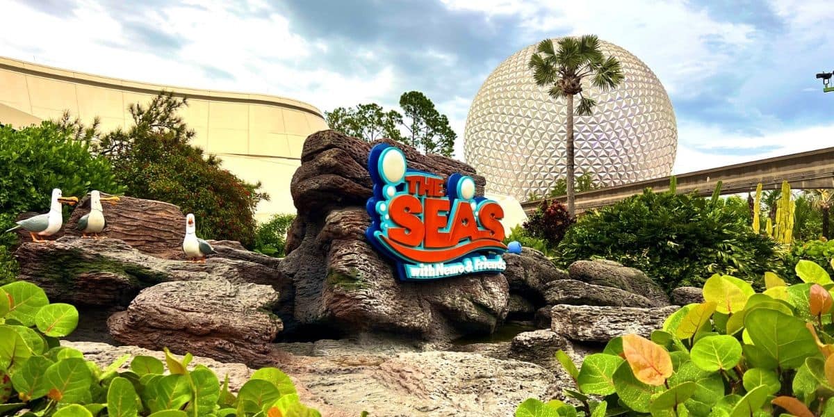 Sign for "The Seas with Nemo & Friends" on rocks surrounded by greenery, near three seagull statues. Epcot's iconic Spaceship Earth dome and Mission: Space can be seen in the background under a partly cloudy sky.