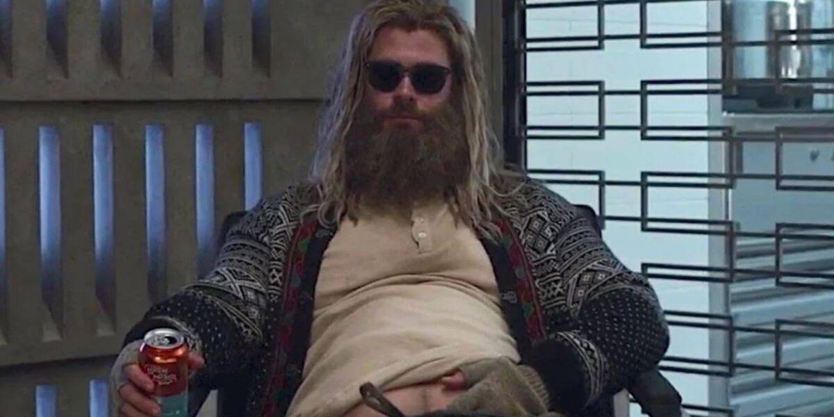 Man with long blond hair and a thick beard, wearing sunglasses and a patterned cardigan, sits slouched in a chair holding a can of soda, exposing his stomach. The modern, industrial setting has a laid-back vibe reminiscent of a Disney character.