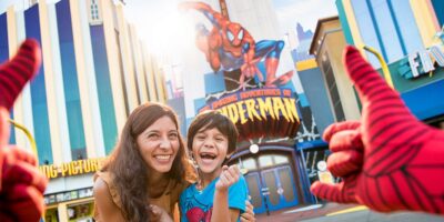 A smiling woman and a young boy pose excitedly in front of The Amazing Adventures of Spider-Man attraction, framed by someone giving a thumbs up with Spider-Man gloves at a vibrant spider-man themed park.