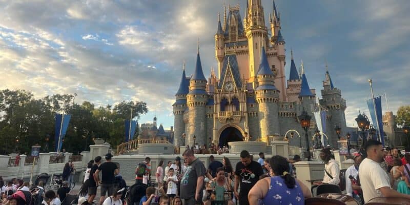 A large crowd of people gathers in front of Cinderella Castle at Disney World, with the castle's spires lit by the setting sun against a partly cloudy sky.