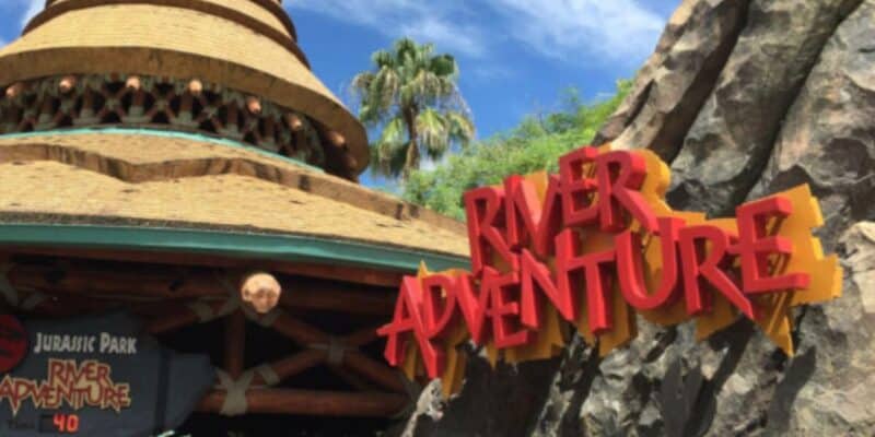 Universal's Jurassic Park River Adventure entrance sign features bold red letters and a themed hut roof, surrounded by rocks and greenery under a blue sky—guiding you to your next attraction.