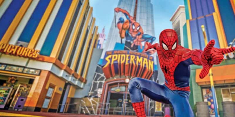 A person in a Spider-Man costume poses in front of the Universal Spider-Man ride, with colorful buildings and a large Spider-Man sign creating a vibrant backdrop.