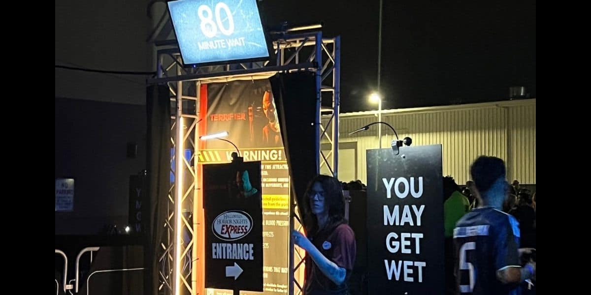 People wait at the dimly lit entrance to a universal attraction at night, with a sign displaying “80 minute wait” and another warning, “You may get wet.” Several visitors are gathered near the entrance.