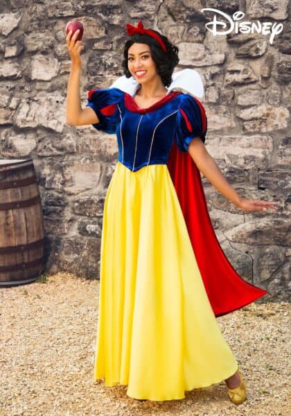 A woman dressed as Snow White in a yellow, blue, and red costume stands outdoors by a stone wall, smiling and holding an apple up in one hand—showcasing one of the best Disney costumes. The Disney logo appears in the top right corner.