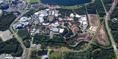 Aerial view of a large theme park complex surrounded by trees, roads, and parking lots, with various buildings, rides, and water areas visible within the park.