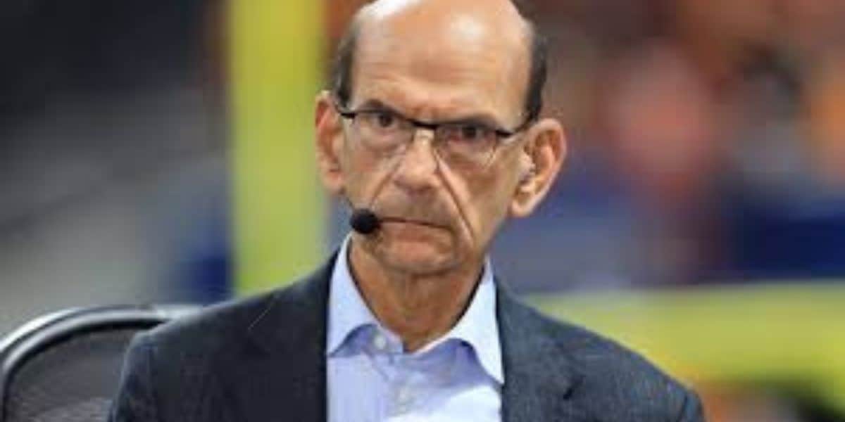 A middle-aged man with glasses, wearing a suit jacket and collared shirt, sits indoors with a serious expression and a headset microphone on.