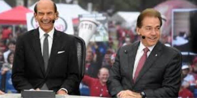 Two men in suits sit at a desk outdoors, smiling and speaking. One wears a black suit and tie, the other a dark suit with a red tie. A crowd with red clothing and signs is visible in the blurred background.