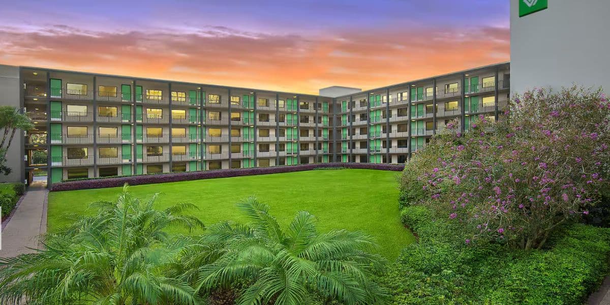 A modern four-story hotel apartment building surrounds a rectangular green lawn with palm trees and flowering shrubs, set against a vibrant orange and purple sunset sky.
