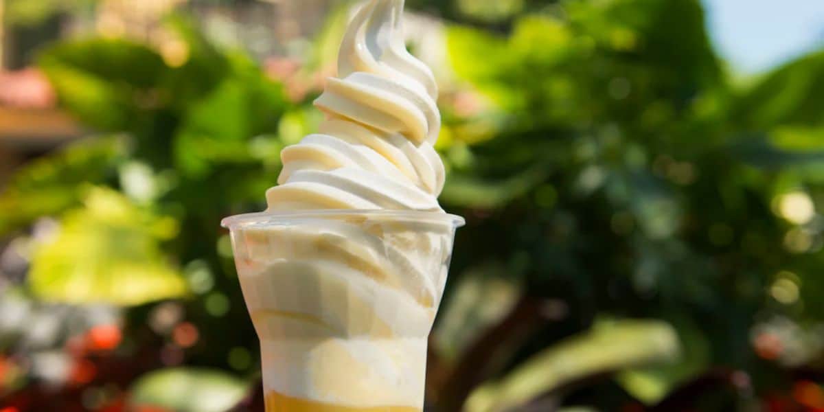 A close-up of a vanilla soft serve ice cream in a clear plastic cup, with yellow sauce at the bottom—reminiscent of a Disney park treat—held outdoors against a blurred background of green foliage.