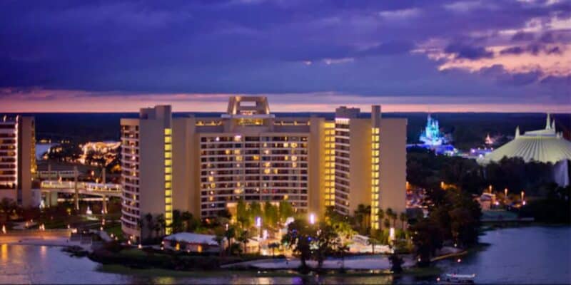 A lit-up modern hotel building stands by the water at dusk, with a dramatic purple and blue sky overhead. Nearby hotels and a theme park with illuminated attractions are visible in the background.