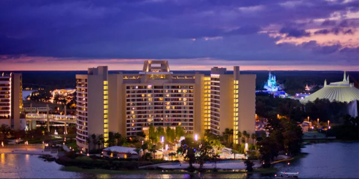 A lit-up modern hotel building stands by the water at dusk, with a dramatic purple and blue sky overhead. Nearby hotels and a theme park with illuminated attractions are visible in the background.
