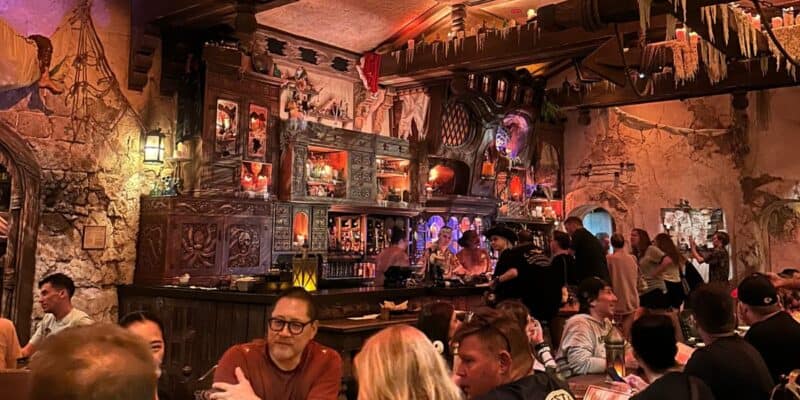 A busy, dimly lit bar with detailed, medieval-style wooden decor. Patrons sit at tables and the bar, surrounded by ornate shelves filled with bottles and skulls, under a warm, ambient glow.