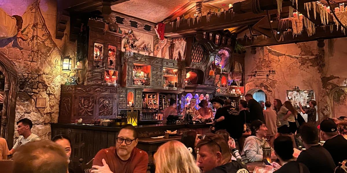 A busy, dimly lit bar with detailed, medieval-style wooden decor. Patrons sit at tables and the bar, surrounded by ornate shelves filled with bottles and skulls, under a warm, ambient glow.