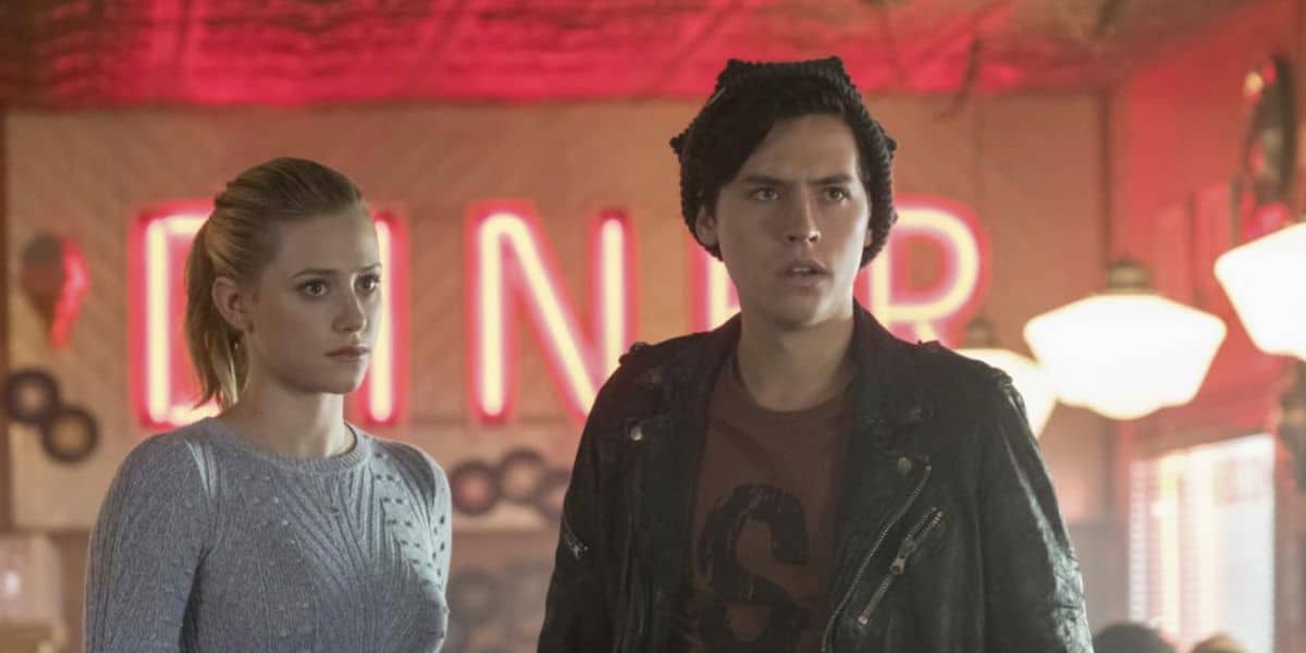 A young woman in a light blue sweater and a young man in a black jacket and beanie stand together in a retro Riverdale-style diner, with a neon "DINER" sign glowing red in the background. Both look serious and concerned.