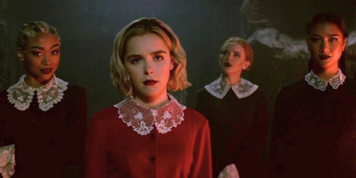 Four young women stand together in gothic attire with lace collars, evoking a Riverdale vibe; the woman in front wears a red dress, while the three behind her wear black dresses, set against a dark, moody background.