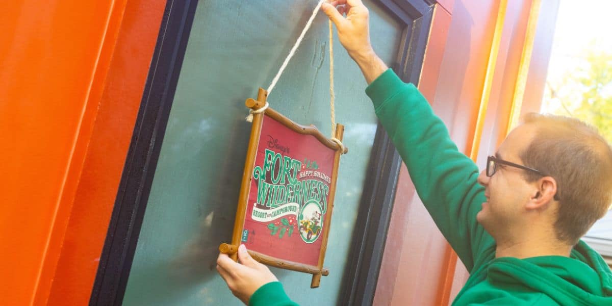 A person in a green hoodie hangs a festive holiday sign reading “Fort Wilderness” on a door with frosted glass, next to an orange wall.