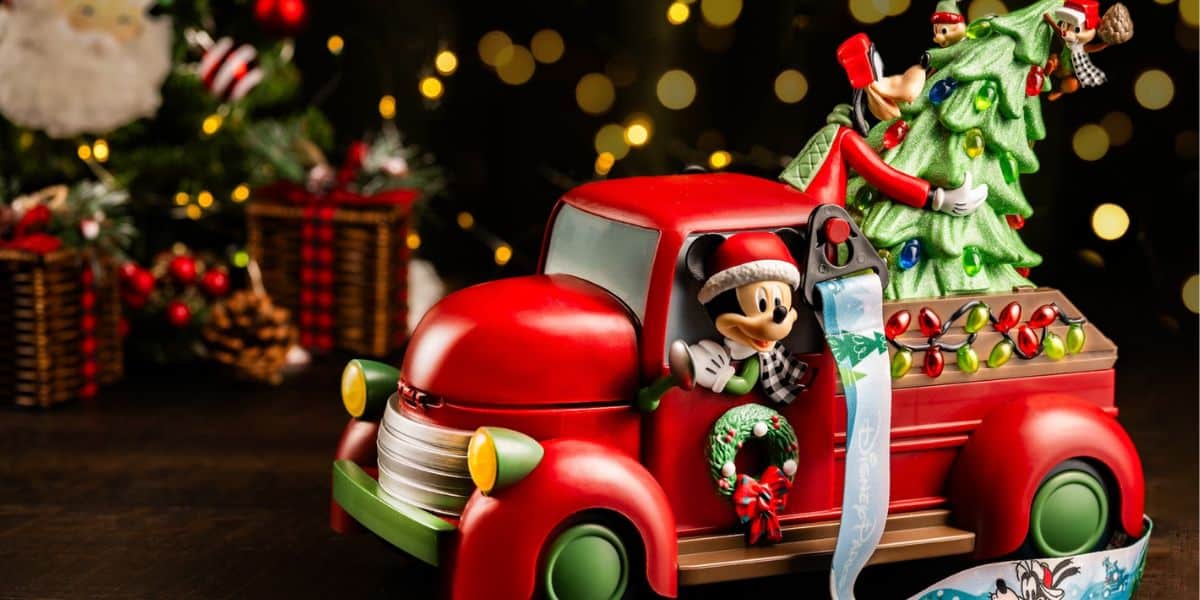A festive red toy truck with Mickey Mouse driving and Minnie Mouse and Pluto decorating a Christmas tree in the truck bed, brings the magic of Disney World to life. Holiday lights and ornaments shine, with a decorated tree glowing in the blurred background.