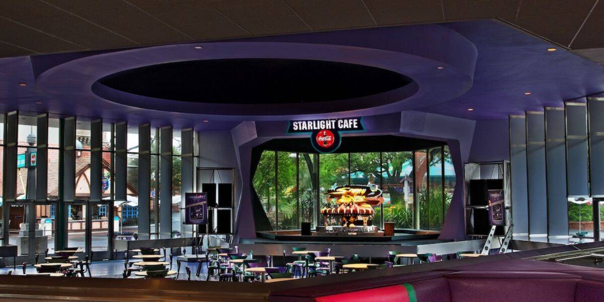 A futuristic café with purple walls, labeled "Starlight Café," features a central stage, Disney-themed décor, glass windows with outdoor views, colorful tables and chairs, and a Coca-Cola sign above the stage.