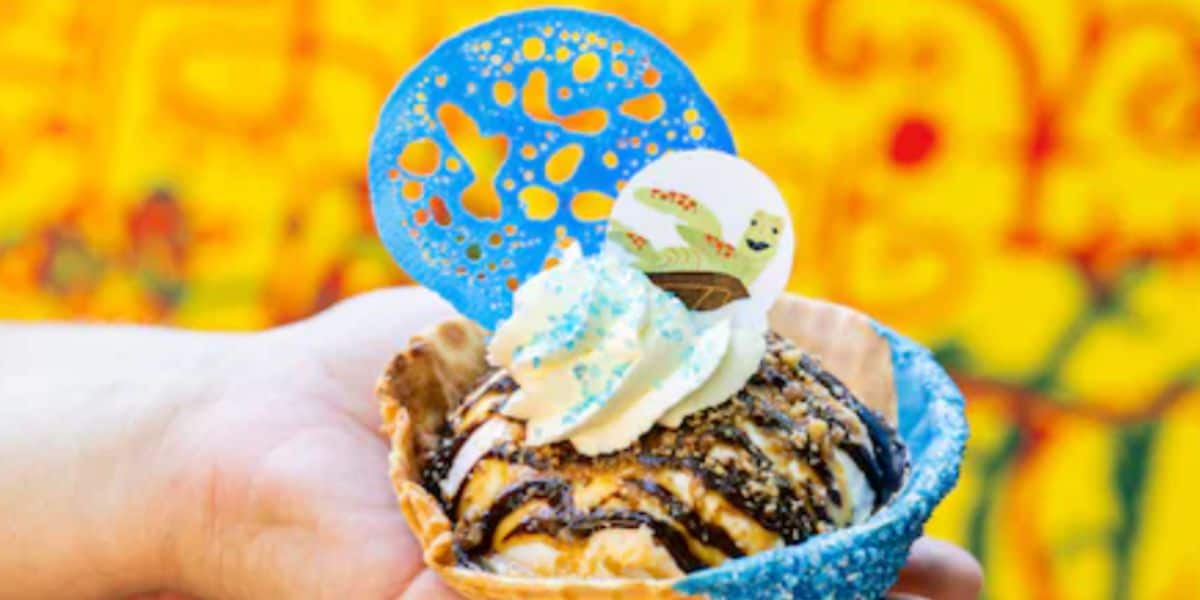 A passholder’s hand holds a waffle bowl filled with ice cream, chocolate syrup, and whipped cream, topped with blue sugar decorations and a white disc featuring cartoon characters, set against a vibrant yellow and orange background.