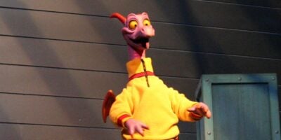 A purple cartoon dragon with orange wings, wearing a yellow sweater, stands in front of a gray paneled wall and a blue prop box, smiling with wide yellow eyes in true Disney style.